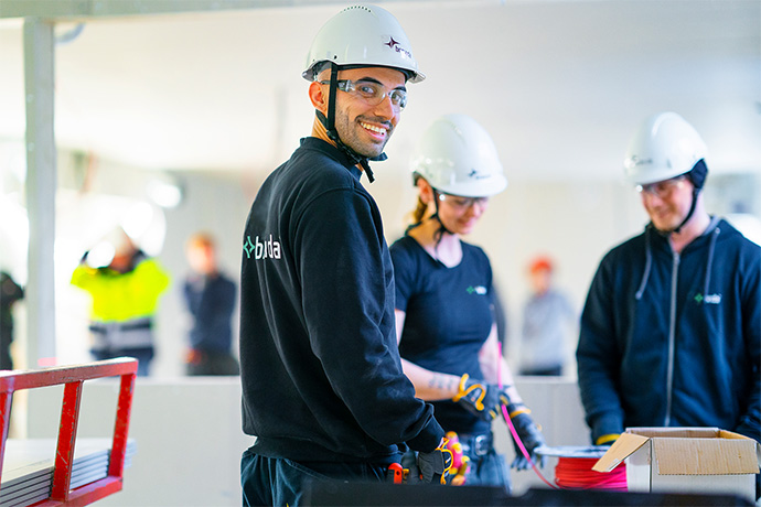Bilden visar en grupp personer i en industriell miljö. Personerna bär skyddshjälmar, skyddshandskar och skyddsglasögon