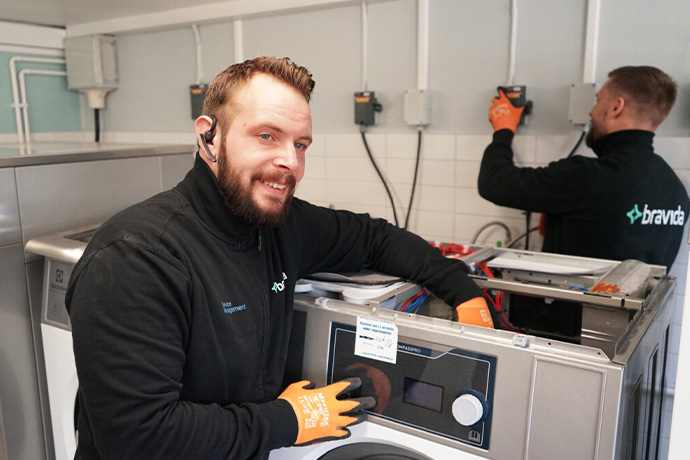 Bilden visar två servicetekniker som arbetar med att installera eller reparera tvättmaskiner. De bär svarta arbetskläder och orangea skyddshandskar. 