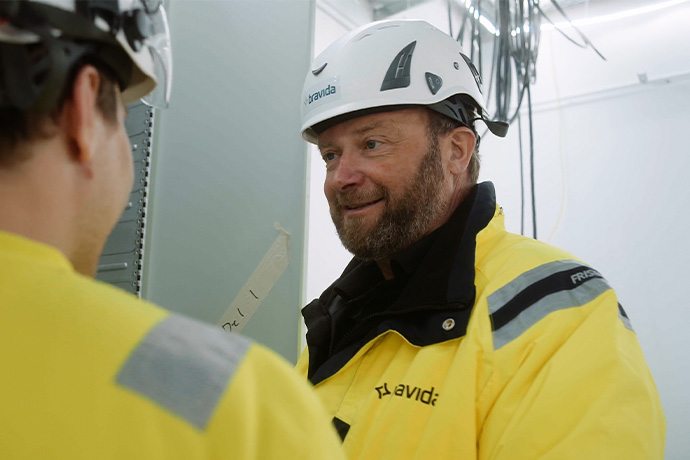 Bilden visar två personer i en arbetsmiljö. Båda bär:  Skyddshjälmar och gula varseljackor.